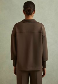 Felpa oversize marrone con colletto e spalle scese, con polsini e orlo a costine. Texture morbida con minimalismo nei dettagli.