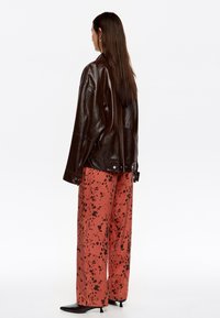 Chaqueta de cuero marrón con un acabado brillante, combinada con pantalones anchos rojos que presentan un patrón moteado en negro. El calzado incluye tacones negros puntiagudos.