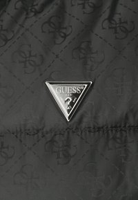 Tessuto trapuntato nero con un motivo monogramma sottile. Una targhetta triangolare in argento con il logo "GUESS" è ben visibile.