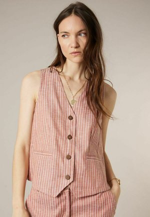 Vrouw met lang bruin haar, gekleed in een mouwloos roze-wit gestreept vest met knopen en bijpassende broek, met een gouden ketting en armband.