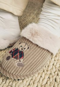 Beige corduroy slippers with a plush fur cuff, featuring an embroidered teddy bear in a patterned sweater on the front. Textured woven floor.