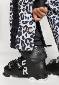 Bocanc de schi negru cu accente albe și pantalon de schi cu imprimeu leopard, având deschidere cu fermoar. Materialul este textil sintetic, cu textură și detalii cu logo.