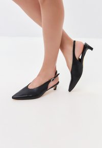 Zapatos de tacón alto de piel negra con diseño slingback, puntera afilada, tacón delgado y detalles metálicos plateados. Textura suave con un diseño de espalda abierta.