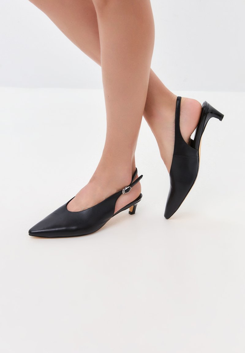 Zapatos de tacón alto de piel negra con diseño slingback, puntera afilada, tacón delgado y detalles metálicos plateados. Textura suave con un diseño de espalda abierta.