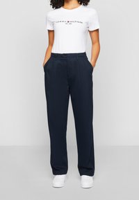 Młoda osoba ubrana w białą koszulkę z logo Tommy Hilfiger, granatowe spodnie z wysokim stanem, z rękami w kieszeniach oraz białe trampki.