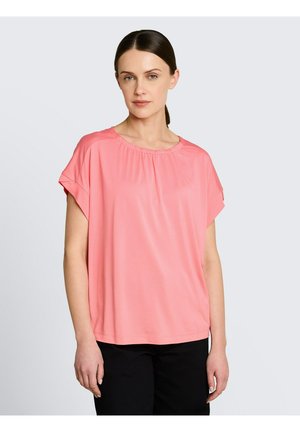 Kobieta o ciemnych włosach, ubrana w luźną różową bluzkę z krótkim rękawem i czarne spodnie, stojąca na jednolitym jasnym tle.