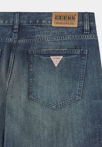 Denim spijkerbroeken in een vervaagde blauwe kleur, met een bruine leren patch en een driehoekig rood-wit logo op de achterzak.