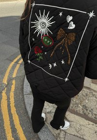 Fekete steppelt dzseki színes hímzett mintákkal: nap, rózsa, masni, szívek, és „LOVE” feliratú folt fehér, zöld és leopárdmintás díszítéssel.