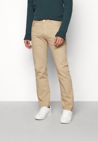Khaki chinos made of cotton, straight-leg design with a button fly, front pockets, and paired with white sneakers and a blue sweater.