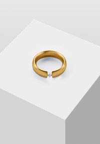 Anello tono oro con una finitura liscia e lucida, caratterizzato da un'apertura e da un diamante tondo e trasparente incastonato al centro. Design semplice e moderno.