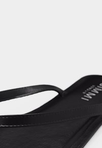 Μαύρο σανδάλι τύπου φλιπ-φλοπ με λεπτές λουρίδες και μερικό λευκό κείμενο λογότυπου ορατό στην περιοχή της φτέρνας, σε απλό ανοιχτό φόντο.