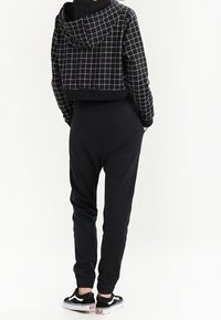 Person som bär en svart hoodie med rutmönster och svarta joggingsbyxor, med svart och vita sneakers, stående med händerna i fickorna.