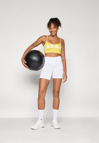 Soutien-gorge de sport jaune avec "NEBBI" en lettres blanches, associé à un short blanc taille haute. Le mannequin tient un ballon de médecine noir. Des sneakers blanches complètent le look.