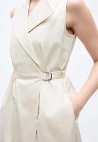 Μπεζ αμάνικο φόρεμα με φαρδιά γιακά και στυλ που τονίζει τη μέση, ασφαλισμένο με ασημένια ζώνη D-ring, το χέρι τοποθετημένο στην πλαϊνή τσέπη.