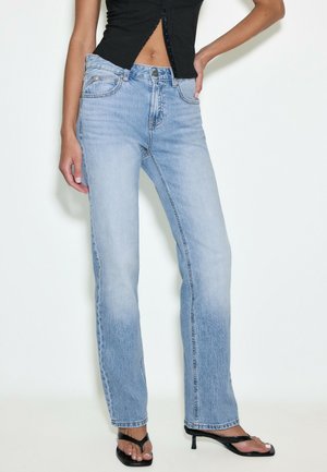 Kvinde iført lyseblå jeans med lige ben, sort cropped top og sorte hælede sandaler, stående mod en ensfarvet lys baggrund.