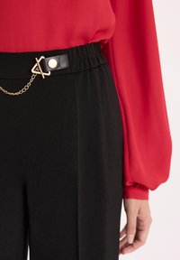 Czarne spodnie z teksturowanej tkaniny, z detalem w postaci złotego łańcuszka i zapięcia na elastycznej talii, w połączeniu z czerwoną bluzką.