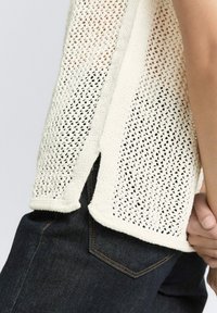 Strickpullover in Creme mit offenem Maschenbild, mit geradem Saum und seitlichen Schlitzen. Kombiniert mit dunkelblauen Jeans aus Denim mit orangefarbener Naht.