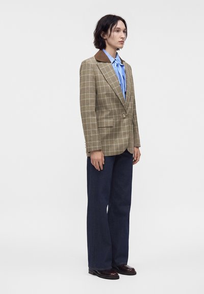 Personne portant un blazer à carreaux marron avec un col marron, un chemisier bleu avec un nœud, un pantalon large foncé et des chaussures marron foncé, debout de côté.