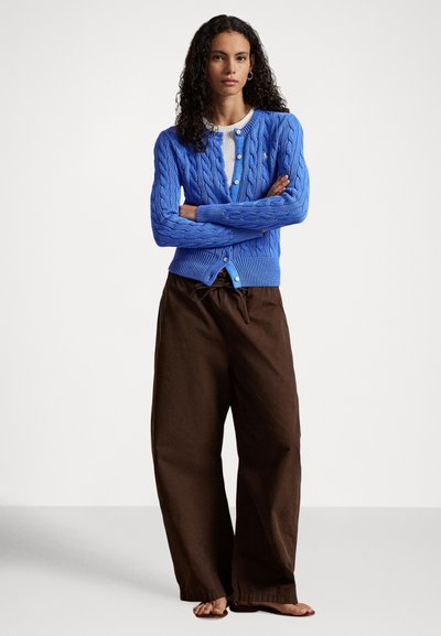 Zila kabeles adījuma kardigans ar apaļu kakla izgriezumu un pogu aizdari, kombinēts ar vaļīgām brūnām biksēm. Apģērbu papildina sandales.