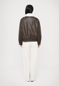 Jachetă bomber maro din piele cu guler de blană artificială albă, mâneci cu manșetă elastică și talie ajustată. Modelul poartă pantaloni albi cu croială largă.