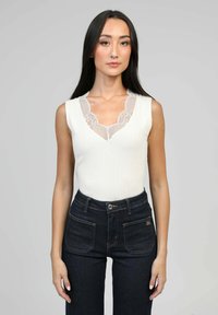 Un haut sans manches blanc avec un décolleté en V et des accents en dentelle, associé à un jeans taille haute en denim foncé avec des poches visibles et des détails de couture.