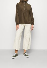 Suéter de pana marrón con cuello alto y cremallera plateada, combinado con pantalones de pierna ancha color crema y zapatillas verdes.