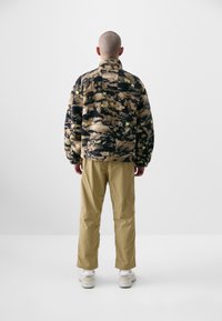 Fleece jacket with a textured, multicolored pattern in brown, black, and green, paired with khaki pants and white sneakers.