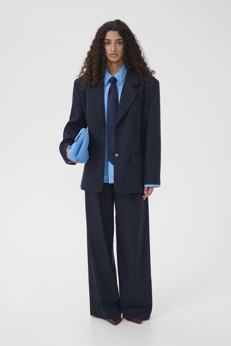 Femme aux cheveux bouclés portant un costume ample bleu marine, une chemise bleu clair, une cravate bleu marine, tenant une pochette bleue duveteuse, debout devant un fond uni.