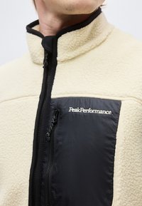 Μπεζ φλις μπουφάν με μαύρη τσέπη στο στήθος με φερμουάρ και λογότυπο "Peak Performance", φορεμένο από άτομο με εμφανές σαγόνι και λαιμό.