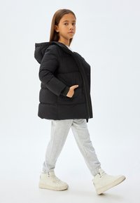 Chaqueta de plumas negra con capucha, cierre de cremallera y puños elásticos. Lleva puesta sobre pantalones de chándal grises y zapatillas altas blancas.