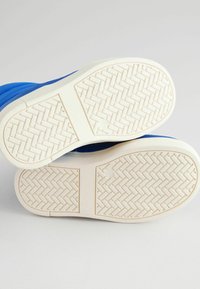Blaue Schuhe mit einer strukturierten weißen Gummisohle. Die Sohle weist ein Fischgrätenmuster und ein kreisförmiges Detail in der Mitte auf.