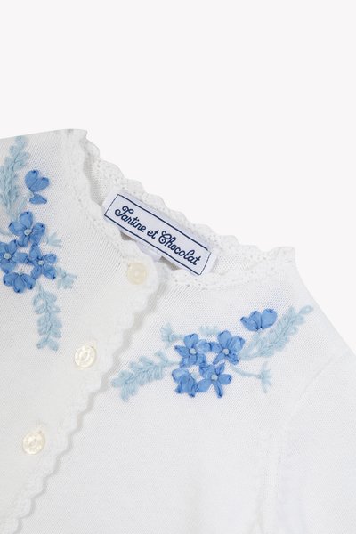 Λευκό ζακέτα με ανοιχτό μπλε κεντημένα λουλούδια, λαιμόκοψη με κυματιστές άκρες και κουμπιά που μοιάζουν μ ε μαργαριτάρια. Η ετικέτα γράφει "Tartine et Chocolat."