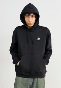 Μαύρη φούτερ με κουκούλα, κατασκευασμένη από μείγμα βαμβακιού, με μπροστινή τσέπη και μικρό λευκό λογότυπο στο αριστερό στήθος. Λείο υφάδι, casual σχέδιο.