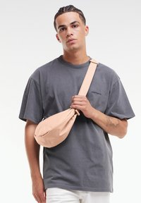 Borsa a tracolla color pesca realizzata in morbido tessuto, forma rotonda e tracolla regolabile, tenuta da una persona che indossa una maglietta grigio scuro.