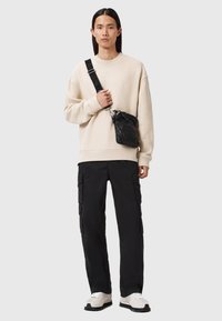 Felpa beige oversize, pantaloni cargo neri con tasche laterali e una piccola borsa a tracolla nera. Sneakers bianche con suole testurizzate.