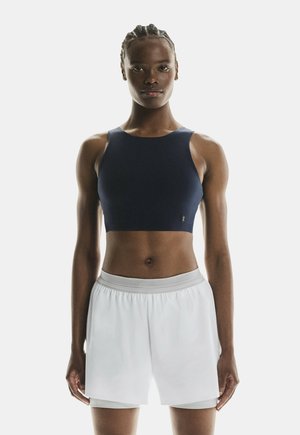 Persona atletica con capelli intrecciati che indossa un crop top blu navy e pantaloncini sportivi bianchi, in piedi su uno sfondo semplice.