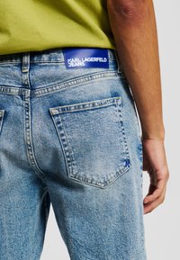 Jeans din denim de culoare albastru deschis, cu un finisaj estompat, prezentând un design al buzunarului din spate și un patch cu logo de culoare albastră pe care scrie "KARL LAGERFELD JEANS."