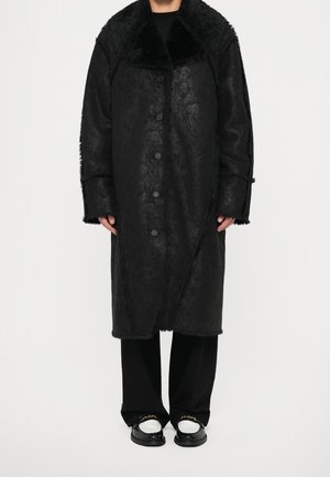 Osoba nosząca długi czarny płaszcz z sztucznego futra z dużym kołnierzem, czarne spodnie i czarno-białe loafersy ze złotym łańcuszkiem.