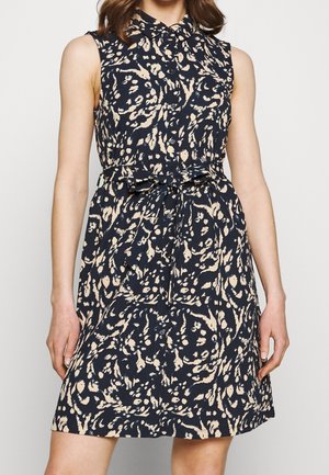 Mouwloze marineblauwe jurk met beige abstract patroon, knoopsluiting aan de voorkant, kraag en een ceintuur die in de taille is geknoopt, gedragen door een staande vrouw.