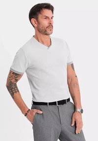 Kortærmet, lysegrå V-hals t-shirt med kontrasterende grå kant ved ærmerne, parret med grå bukser og et sort bælte, båret af en mandlig model.