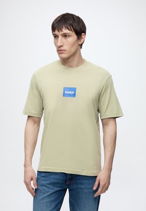 Junger Mann trägt beige HUGO T-Shirt und blaue Jeans, schaut seitlich mit neutralem Gesichtsausdruck vor einfachem Hintergrund.