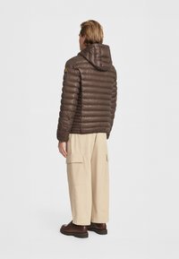 Giacca puffa marrone con cappuccio, caratterizzata da linee orizzontali cucite. Indossata con pantaloni cargo beige, scarpe marroni e tasche laterali visibili.
