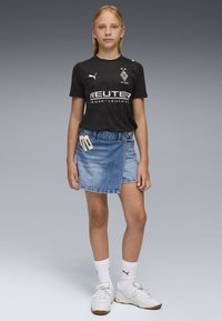 Ragazza in piedi con indosso una maglietta sportiva nera, una gonna di jeans avvolgente con fermagli in perla, calzini bianchi Puma e sneakers bianche su uno sfondo grigio.