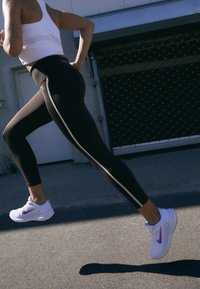 Černé sportovní legíny s bílým bočním pruhem, doplněné bílými a fialovými teniskami, které jsou nošeny při běhu po betonovém povrchu.