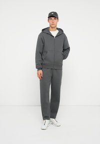 Felpa grigia con zip e tasche a marsupio, abbinata a pantaloni della tuta grigi, indossata con un berretto scuro e sneaker bianche.