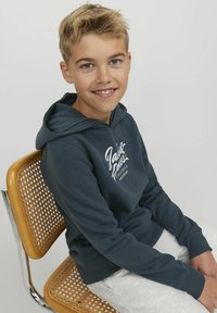 Felpa con cappuccio blu scuro e testo del logo bianco. Materiale misto cotone, tasca a marsupio, polsini a costine, indossata su pantaloni della tuta grigi. Seduto su una sedia.