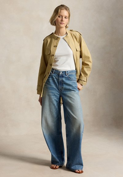 Femeie purtând o jachetă bej cu nasturi peste un maiou alb, blugi albaștri palidați cu pantaloni largi și sandale cu degetele descoperite, stând în picioare pe un fundal simplu.