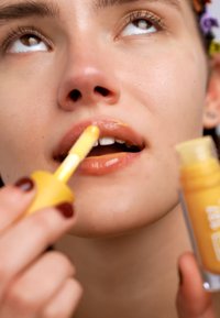 Lucidalabbra in un contenitore trasparente con un tappo giallo. L'applicatore è giallo e il lucidalabbra ha una tonalità arancione calda e lucida.