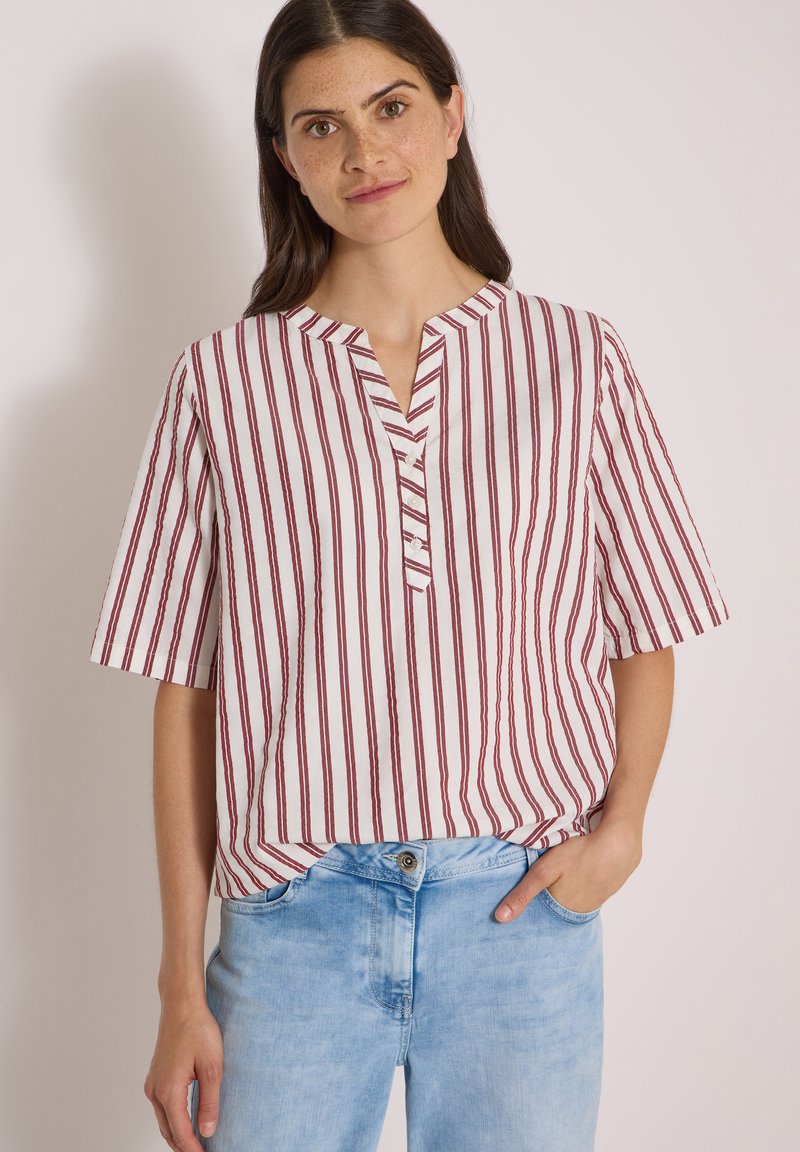 Femme aux cheveux foncés portant une chemise blanche à rayures rouges verticales et un jean bleu clair, debout avec une main dans la poche devant un fond uni.