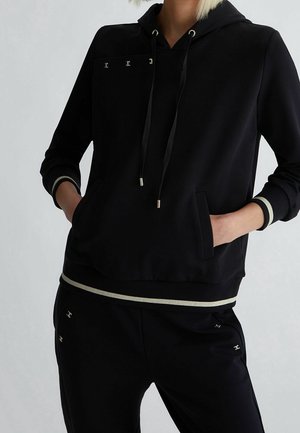 Person trägt schwarzen Kapuzenpullover und passende schwarze Hose mit dezenten silbernen Akzenten und kleinen metallischen Logodetails. Hände in den Taschen des Kapuzenpullovers.